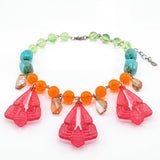 A vibrant statement necklace featuring large, translucent pink Art Deco style teardrop pendants, accented by bright orange beads, turquoise blue beads, and light green beads on a silver-tone chain.